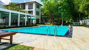 Une piscine avec un banc en bois, un espace de repos couvert et un bâtiment doté de grandes fenêtres et d’un balcon.