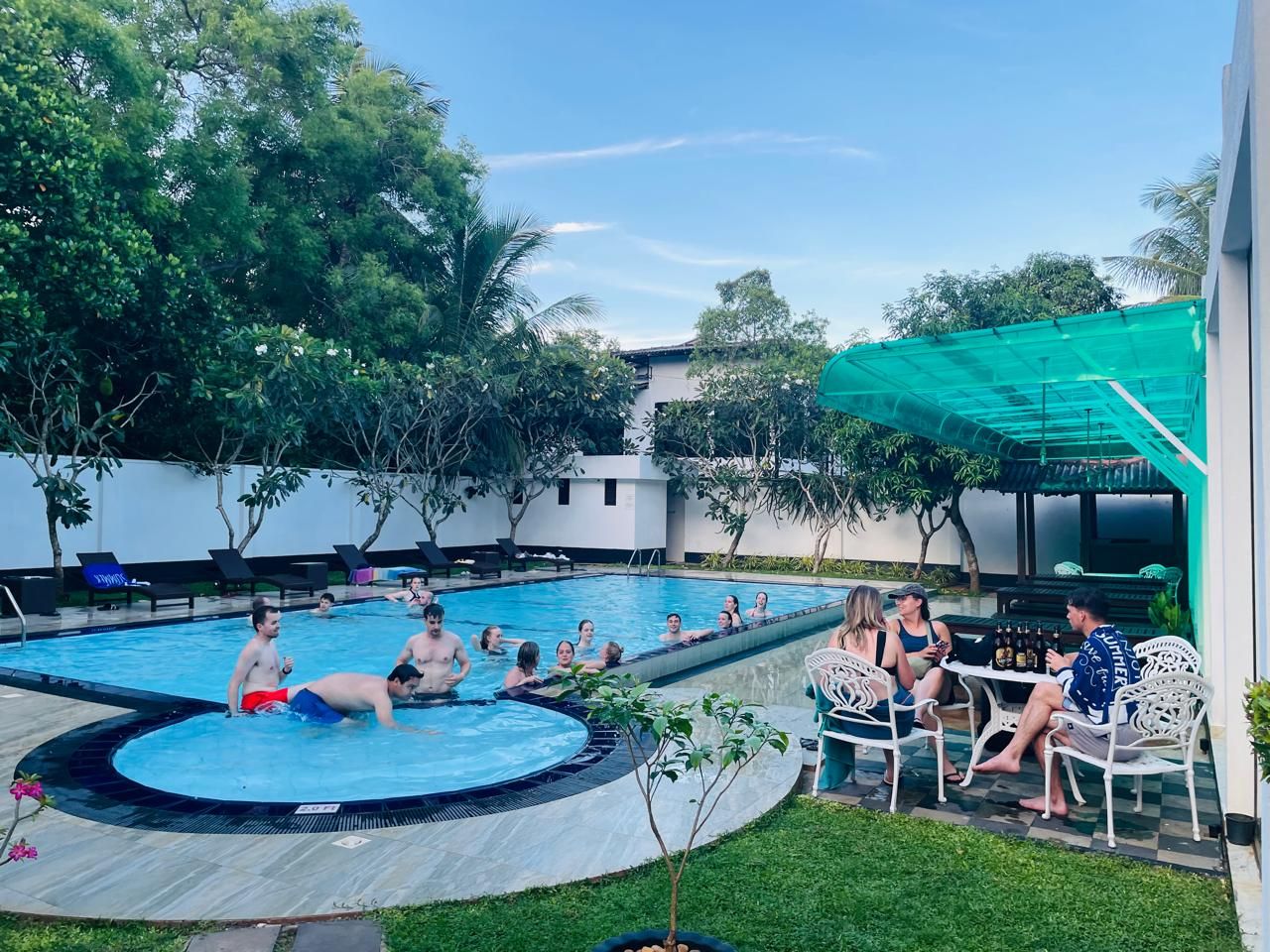 A swimming pool area with people swimming and socializing, surrounded by greenery and outdoor furniture.