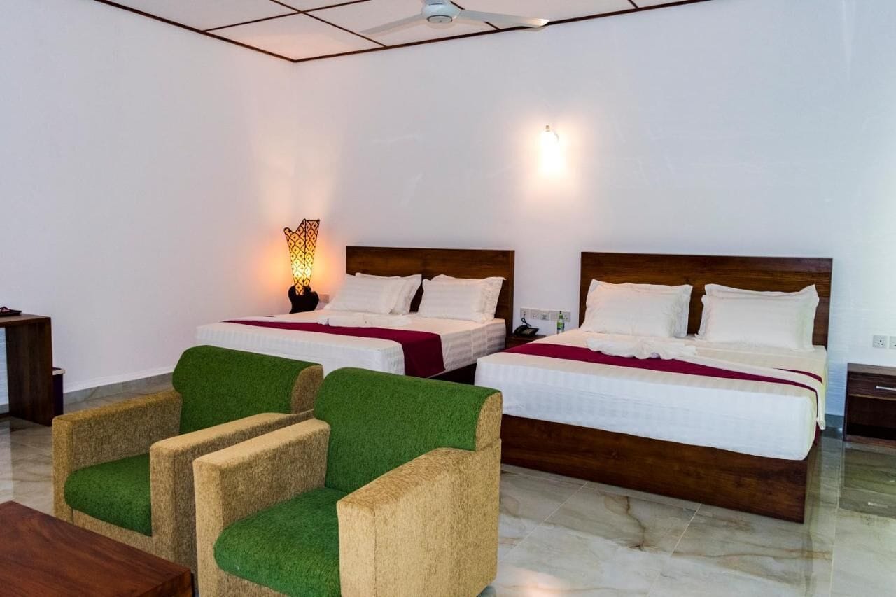 A hotel room with two single beds, a nightstand, a lamp, and green upholstered chairs.