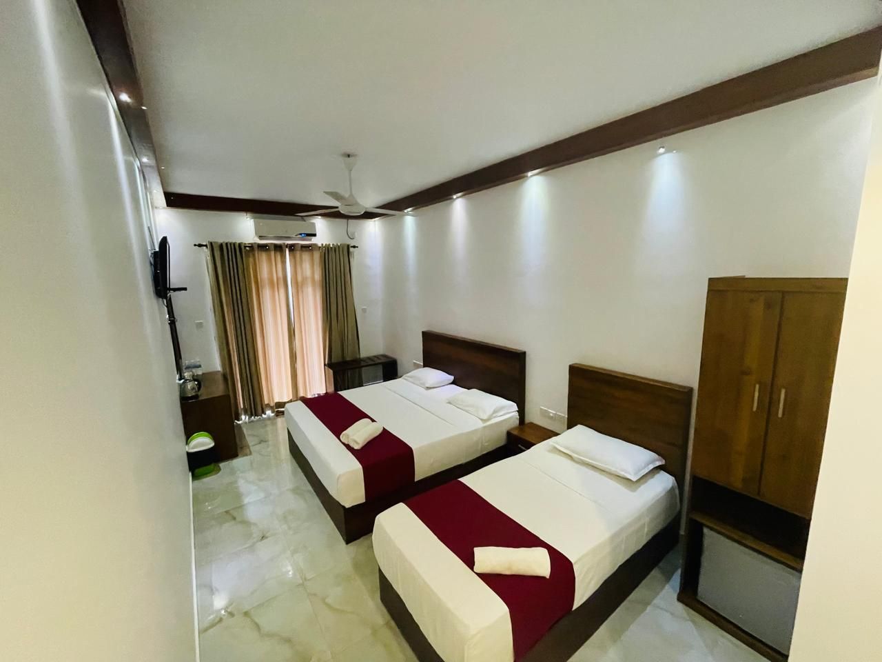 A hotel room with two beds, a wardrobe, a desk, and a ceiling fan.