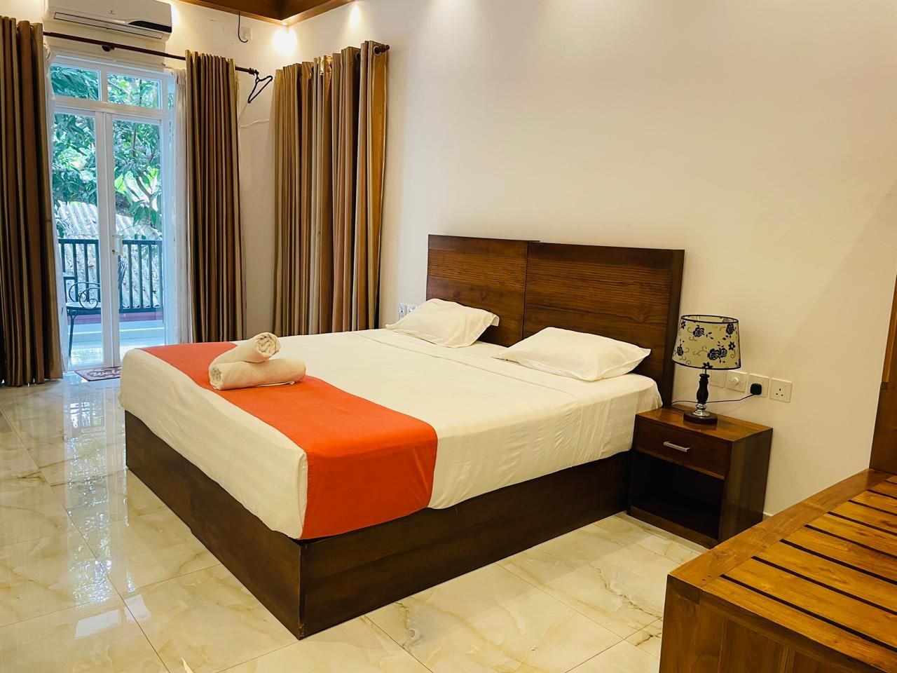 A hotel room with a large bed, a wooden nightstand, a lamp, and a view of the outdoors through a glass door.