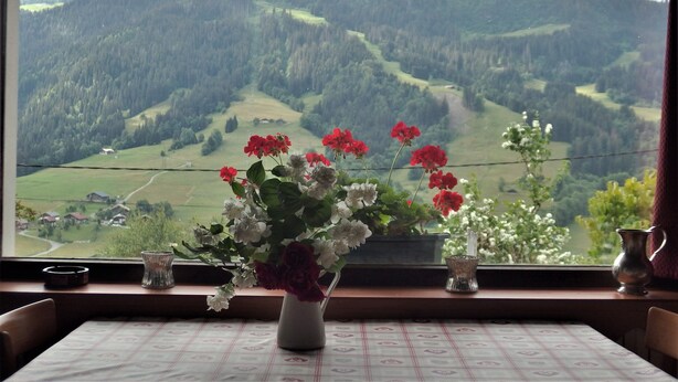 A dining table with a vase of flowers in the foreground and a scenic mountain view through a window in the background. - Coquet Studio (Praz-sur-Arly)