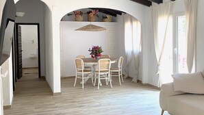 Amplia sala de estar con piso de madera, una mesa redonda de comedor con sillas, un sofá y un banco. Cuenta con vigas de madera en el techo y un ventanal con cortinas.