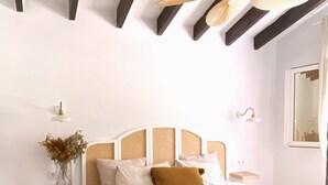 Un dormitorio con cama, ventilador de techo, mesita de noche con lámpara, estante en la pared con un florero, y un ventanal grande.