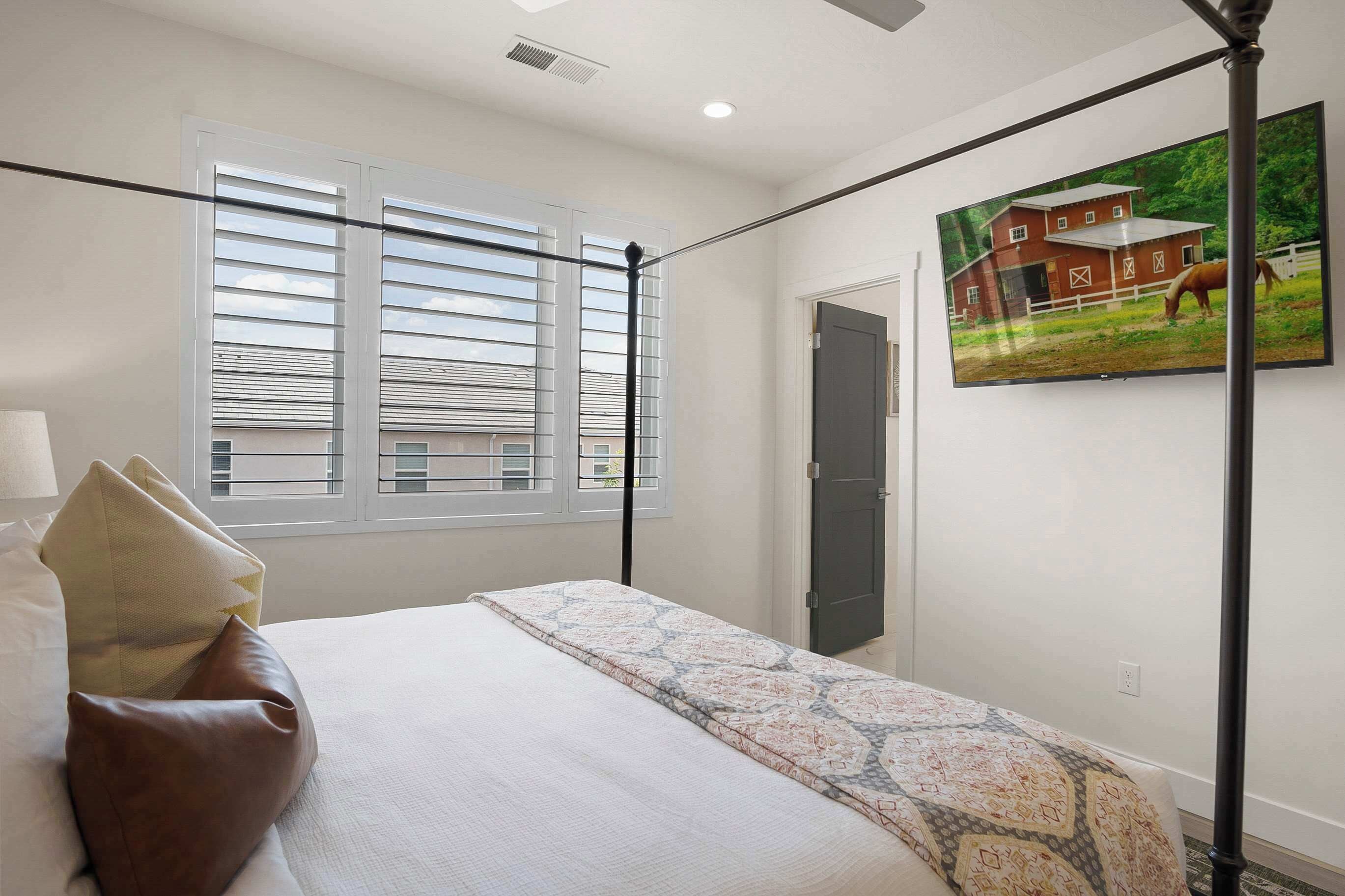 A bedroom with a bed, pillows, a window with blinds, a door, and a wall-mounted TV displaying a rural scene.