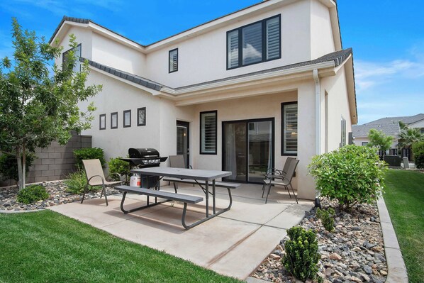 A two-story house with a patio area featuring a grill, a picnic table, and outdoor seating.