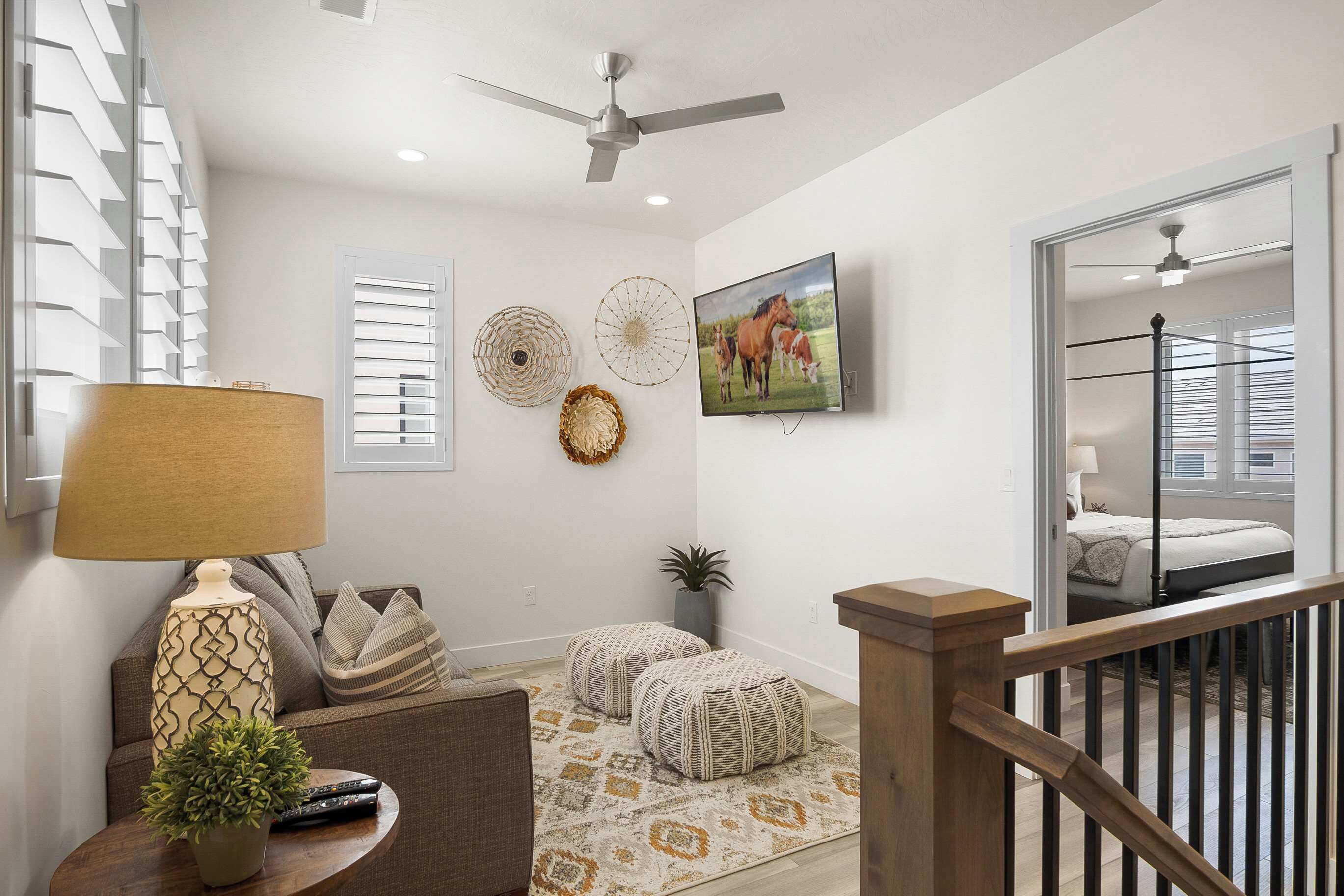 A modern living room with a ceiling fan, a TV mounted on the wall, a sofa, ottomans, a lamp, and a potted plant.