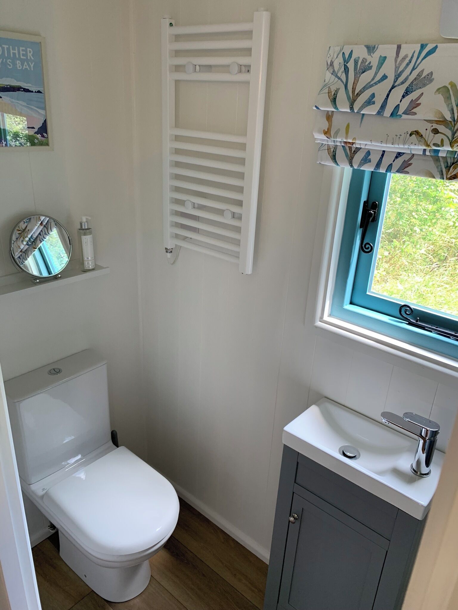 Une petite salle de bain avec des toilettes blanches, un petit lavabo doté d’un robinet chromé, un porte-serviettes et une fenêtre donnant sur un espace verdoyant.