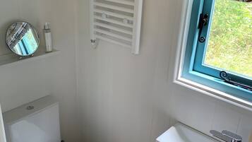 Une petite salle de bain avec des toilettes blanches, un petit lavabo doté d’un robinet chromé, un porte-serviettes et une fenêtre donnant sur un espace verdoyant.