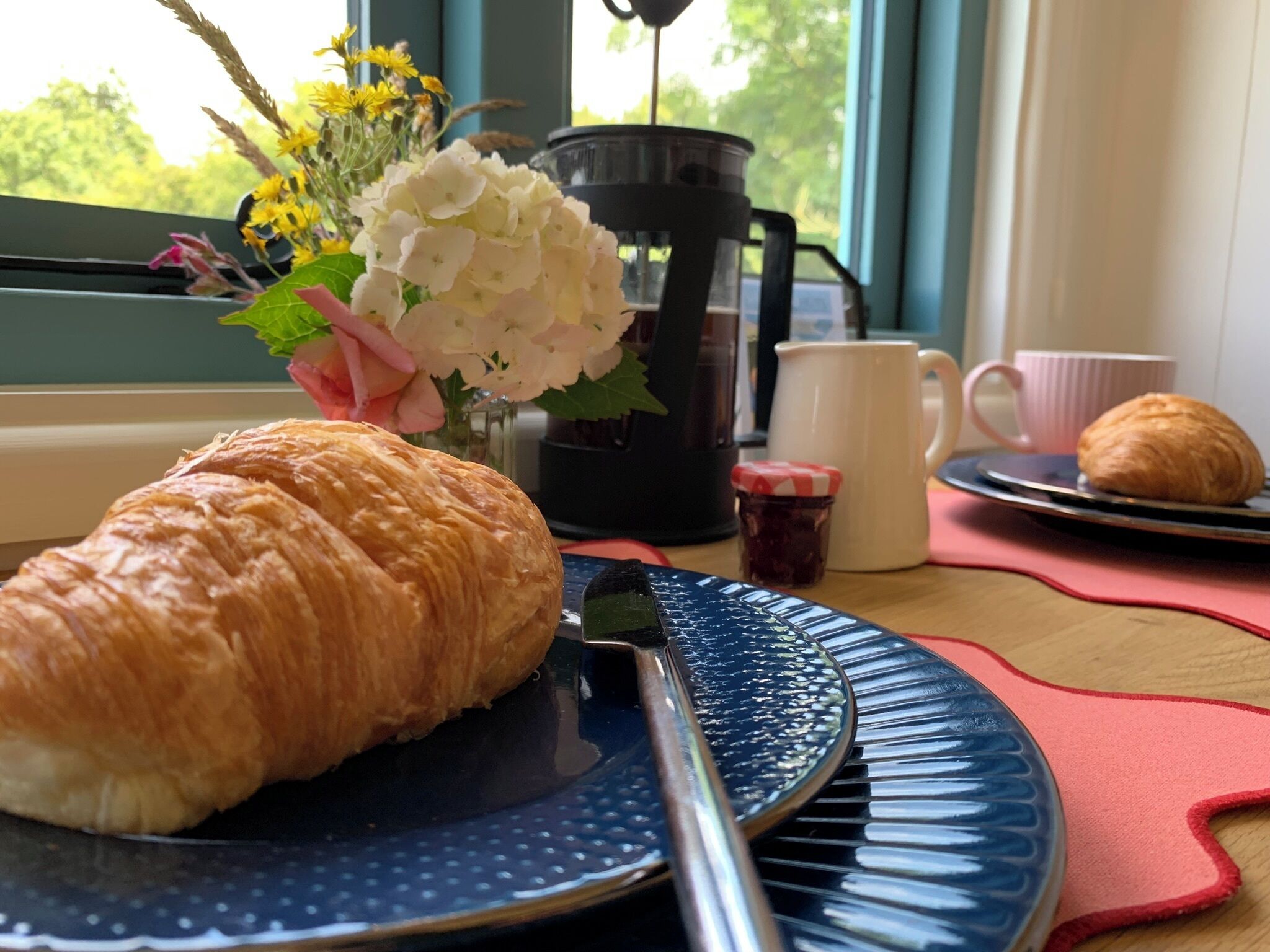 Um croissant num prato, uma cafeteira, uma chávena e um frasco de compota sobre uma mesa com vista para a janela.