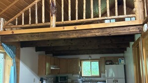 Interior rústico de una cabaña con una terraza de madera, zona de cocina con estufa, microondas y refrigerador, y una mesa de comedor con sillas.