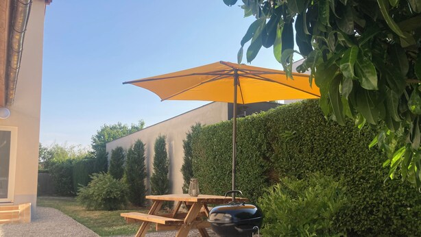 Een terras met een houten tafel, een barbecue en een gele parasol.