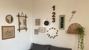 Een woonkamer met een grijze bank, een spiegel, lijstjes met foto’s, een wandklok en decoratieve objecten, waaronder een nummerreeks, een cirkelvormige krans en een plant.