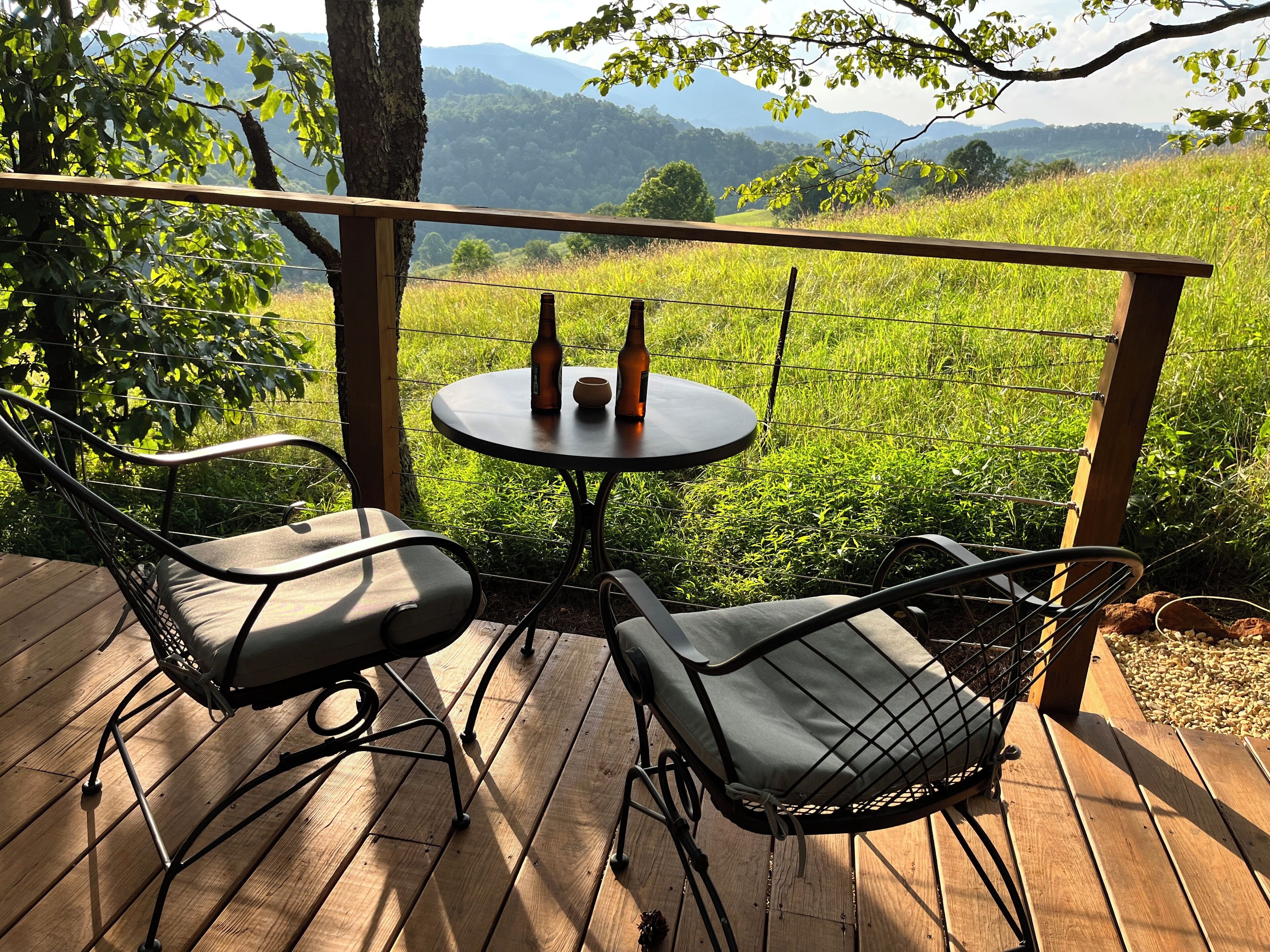 A wooden deck with two metal chairs and a small round table, set against a backdrop of a grassy field and distant mountains.