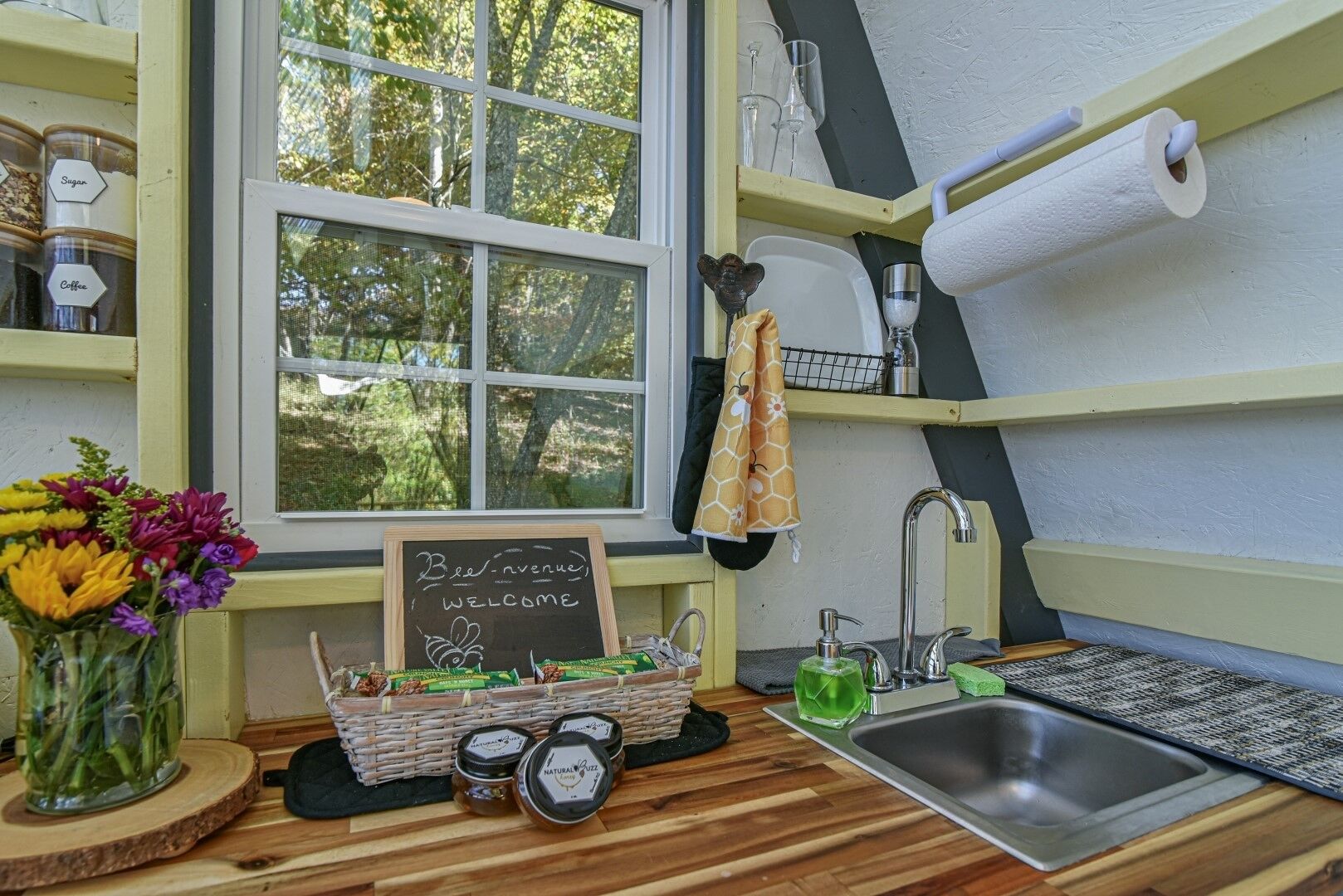 Een keuken met een houten aanrecht, een spoelbak, een prikbord met de tekst 'Bee Avenue Welcome', potten honing en een bloevas.