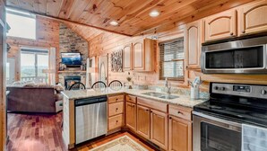A modern kitchen with wooden cabinets, stainless steel appliances, and a stone fireplace. - Mountain Views, Hot Tub, Pool Table, Fire Pit, Dogs Welcome, 3 King Beds (Sevierville)