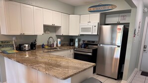 A kitchen with white cabinets, a granite countertop, a stainless steel refrigerator, a microwave, a toaster, and a trash can. - Oceanfront Paradise - Steps to Beach & Rocket Launches! (Cocoa Beach)