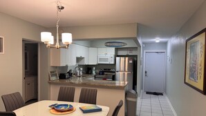 A compact kitchen with white cabinets, a stainless steel refrigerator, and a microwave. There's a dining area with a white table and chairs, and a trash can is visible. - Oceanfront Paradise - Steps to Beach & Rocket Launches! (Cocoa Beach)