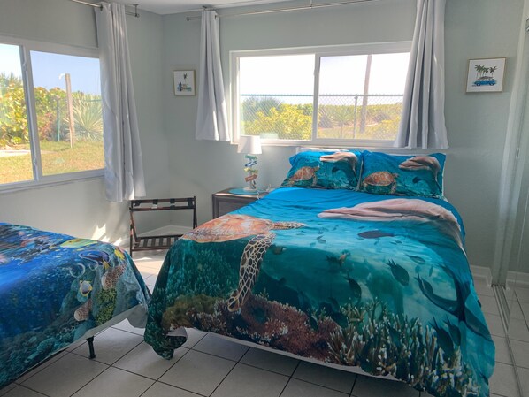 A bedroom with a bed featuring a marine-themed bedspread, a bedside table with a lamp, and a view of the outdoors through a window. - Oceanfront Paradise - Steps to Beach & Rocket Launches! (Cocoa Beach)