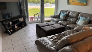 A living room with a leather sofa, ottoman, and a flat-screen TV. - Oceanfront Paradise - Steps to Beach & Rocket Launches! (Cocoa Beach)