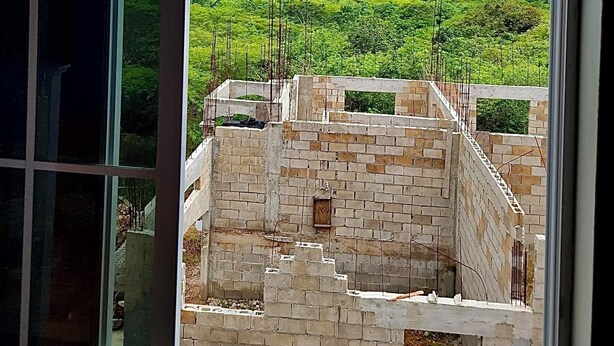 A view through a window of a building under construction with a clear sky and green landscape in the background. - Beautiful Top Apartment in St. Elizabeth, Jamaica with an amazing Mountain View! (Junction)