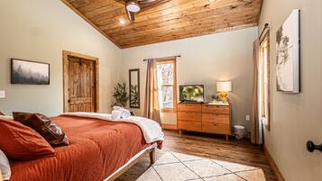 Une chambre avec un plafond en bois, un lit recouvert d’une courtepointe rouge, une commode, un téléviseur et une fenêtre avec des rideaux.