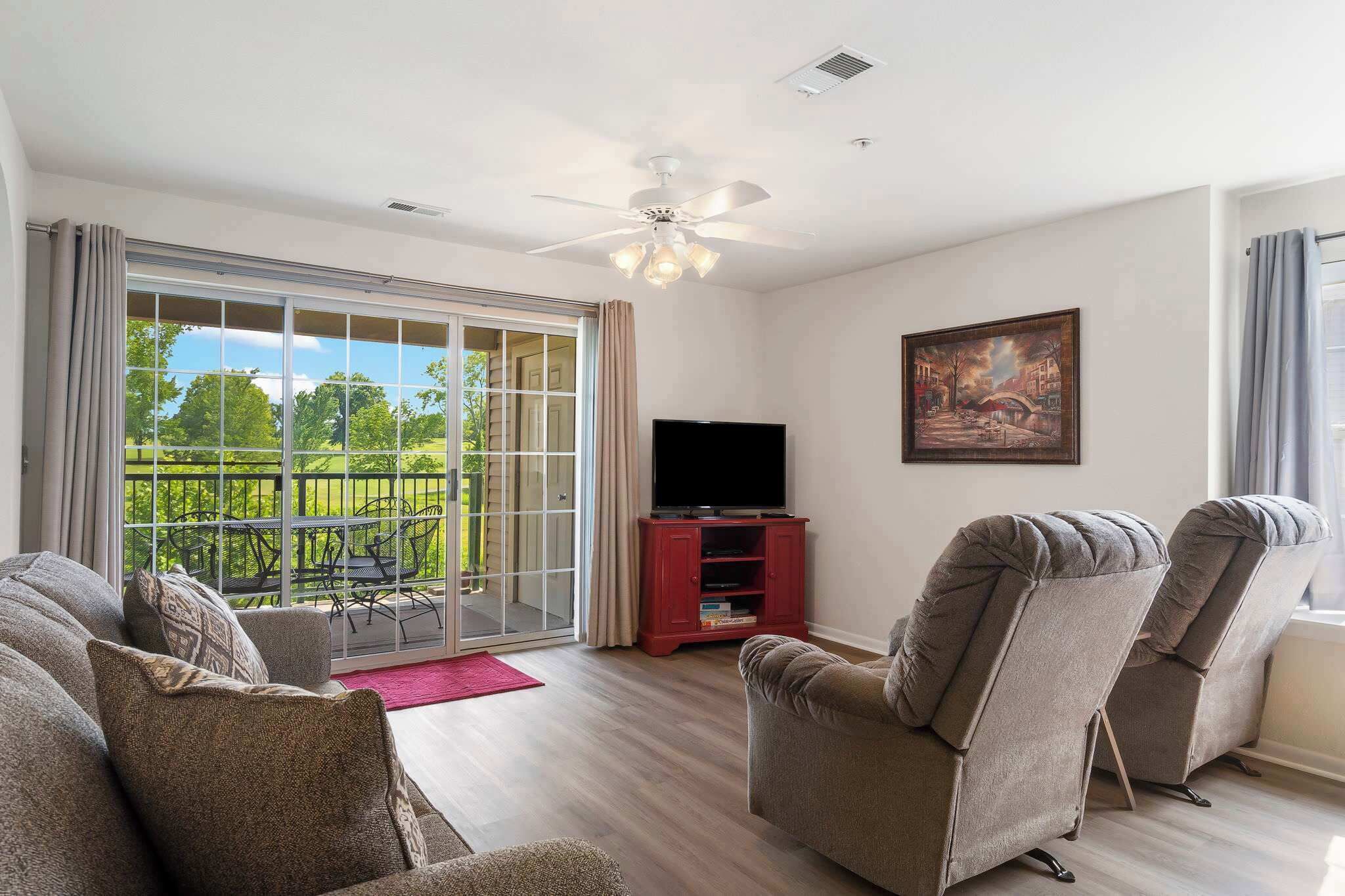 A living room with a sliding glass door leading to a balcony, a ceiling fan, a flat-screen TV on a red stand, and two grey armchairs.