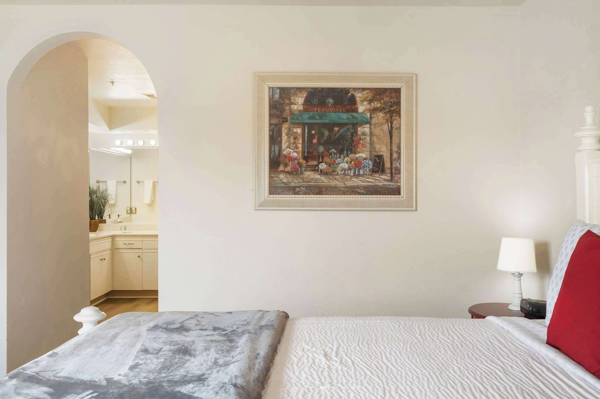 A bedroom with a bed, a nightstand, a lamp, and a framed picture of a flower shop on the wall.