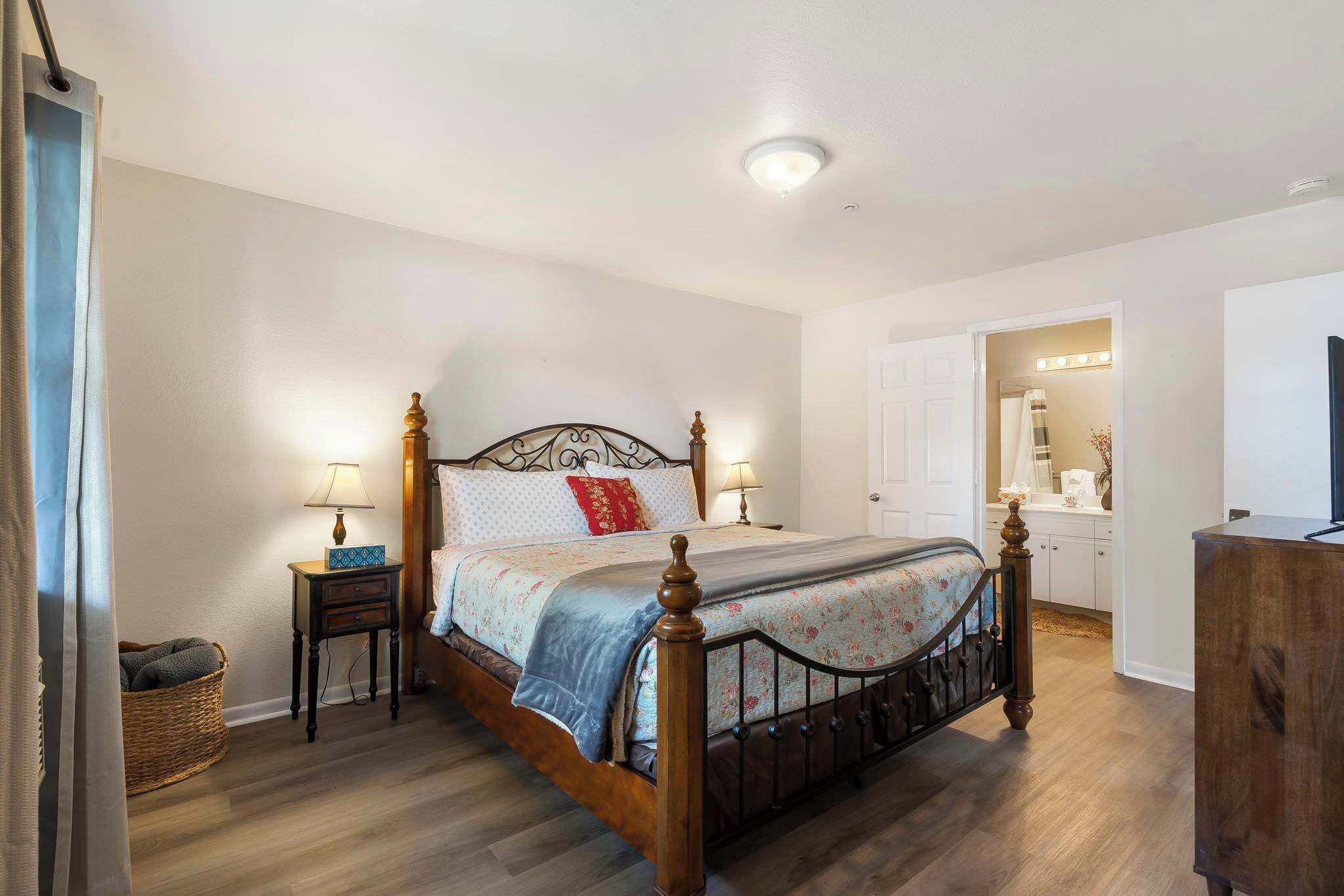 A bedroom with a wooden bed, two bedside tables with lamps, a wicker basket, and a door leading to a bathroom with a mirror and lights.