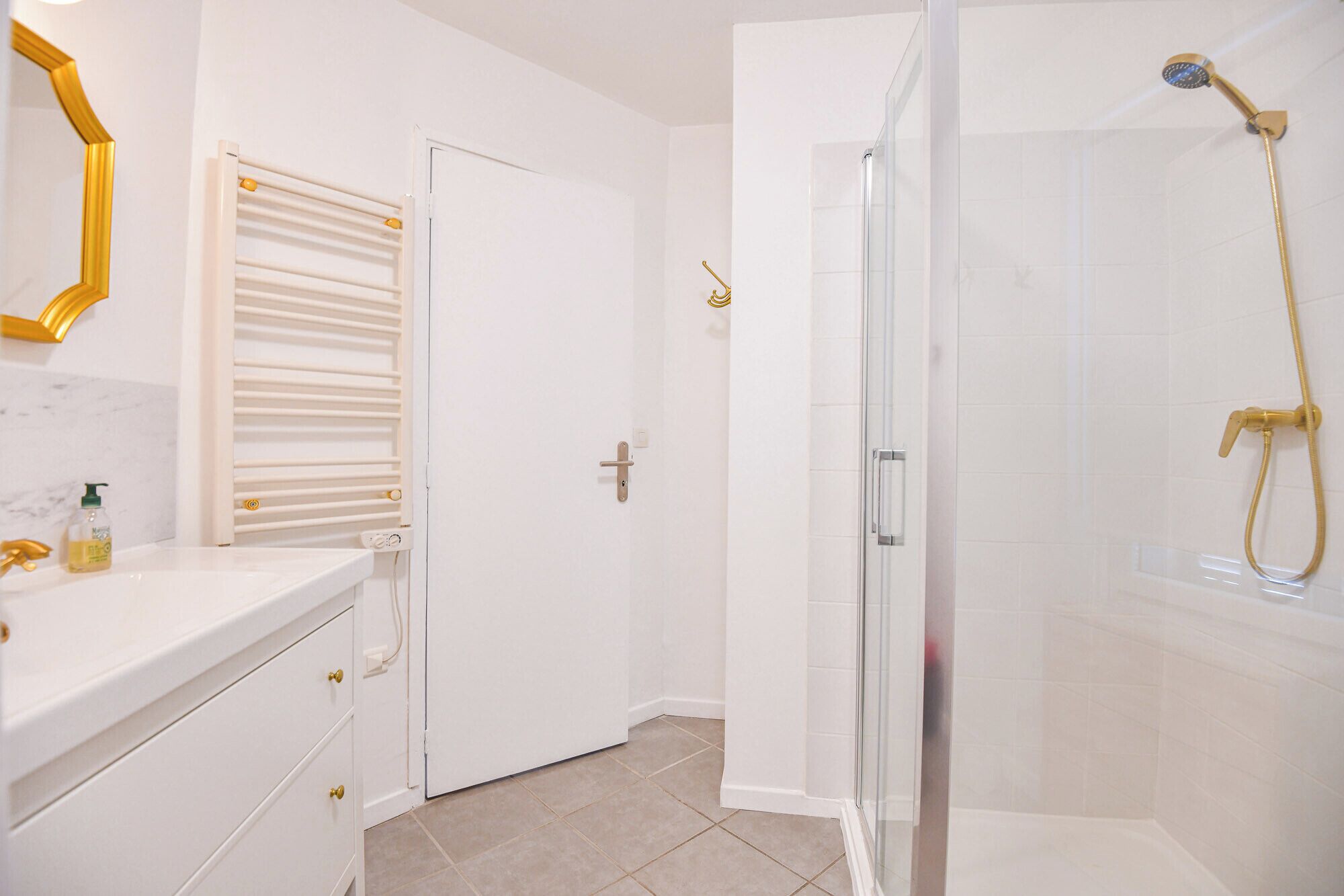 Une salle de bain moderne avec une douche à l’italienne en verre, un meuble-lavabo blanc avec un robinet doré et une porte dotée d’un radiateur blanc.
