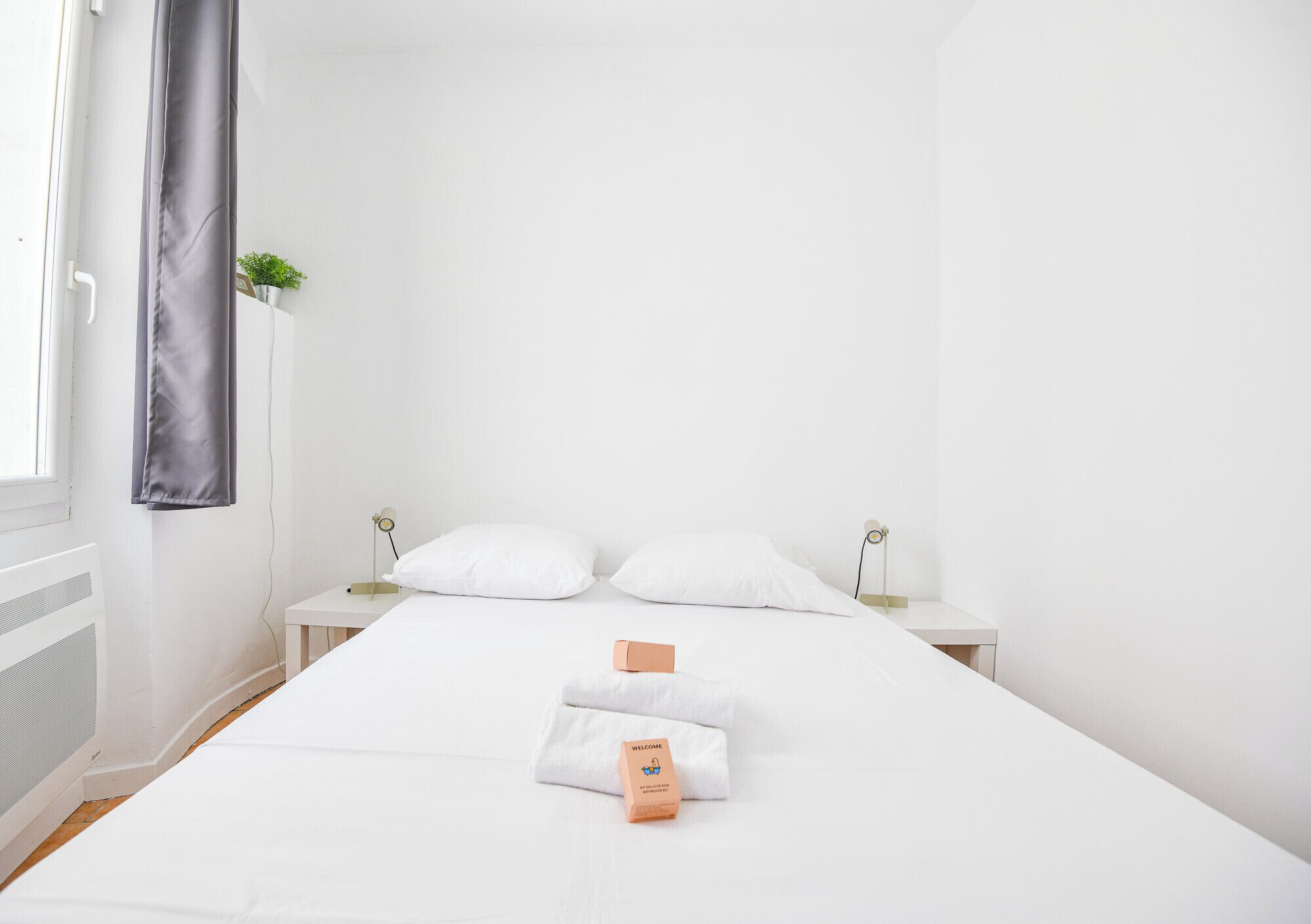 Une chambre d’hôtel propre et blanche, avec un lit bien fait, une table de chevet avec une lampe et une petite plante posée sur la table de chevet.