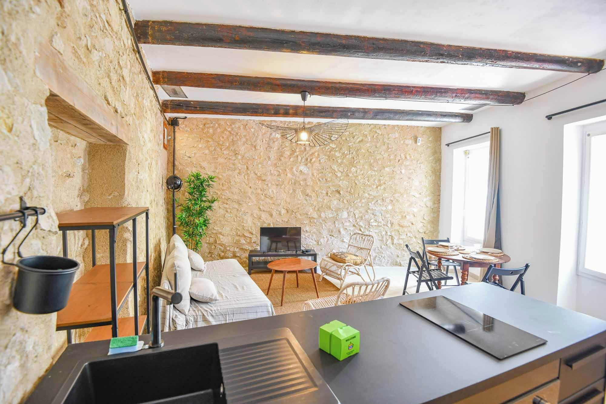 A room with a stone wall, a bed, a desk with a laptop, a dining table with chairs, and a hanging light fixture.