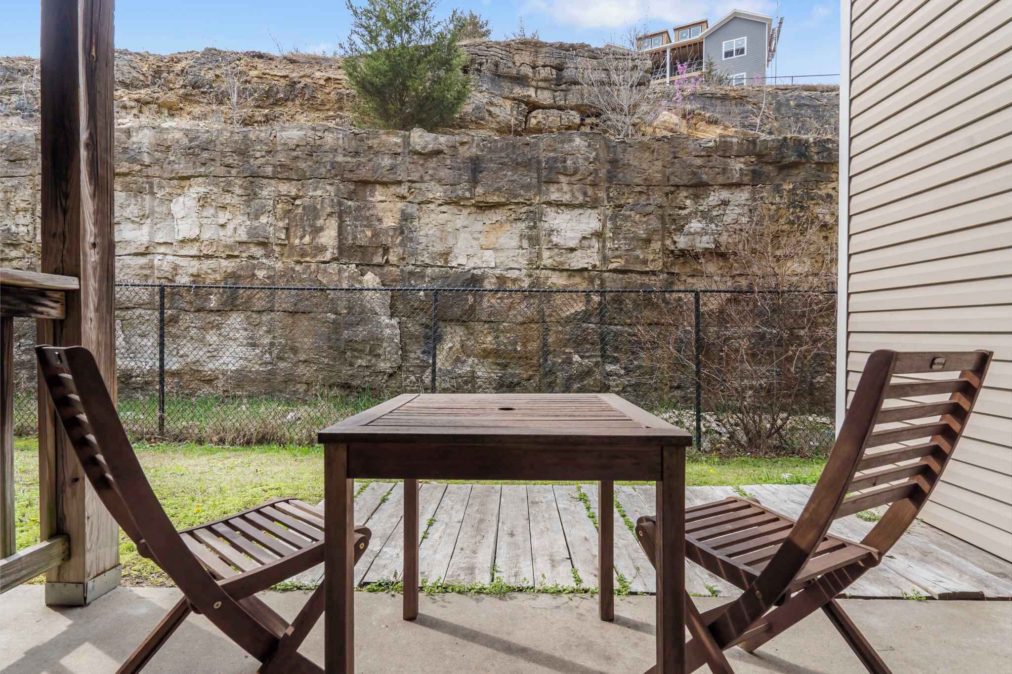 A patio with a wooden table and two chairs, a stone wall, and a house in the background.