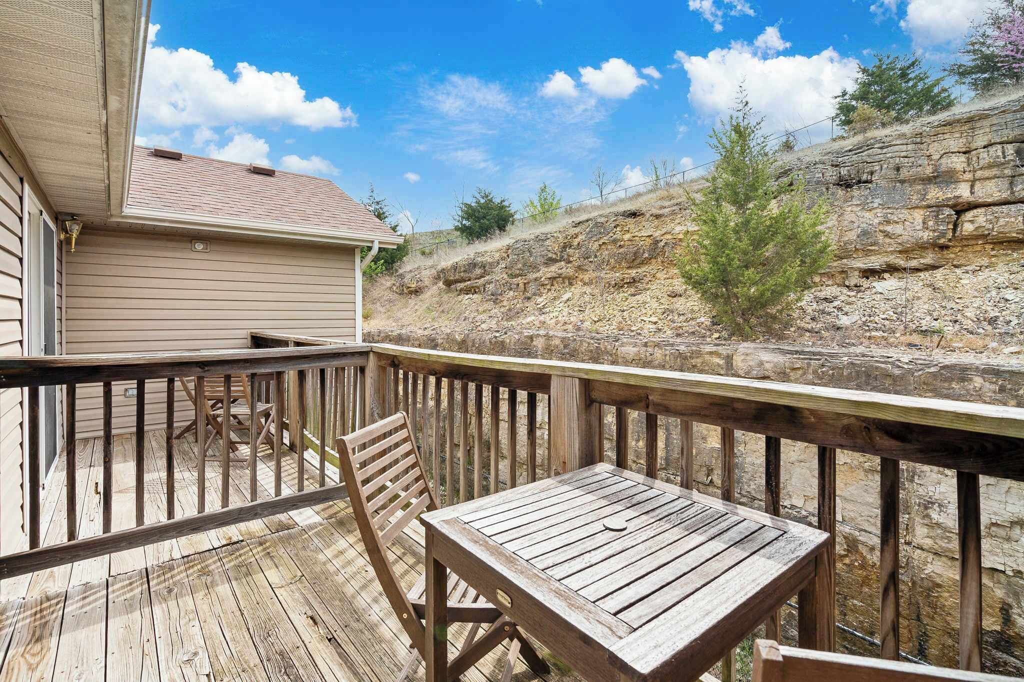 A wooden deck with a table and chairs, overlooking a rocky hillside and a house.