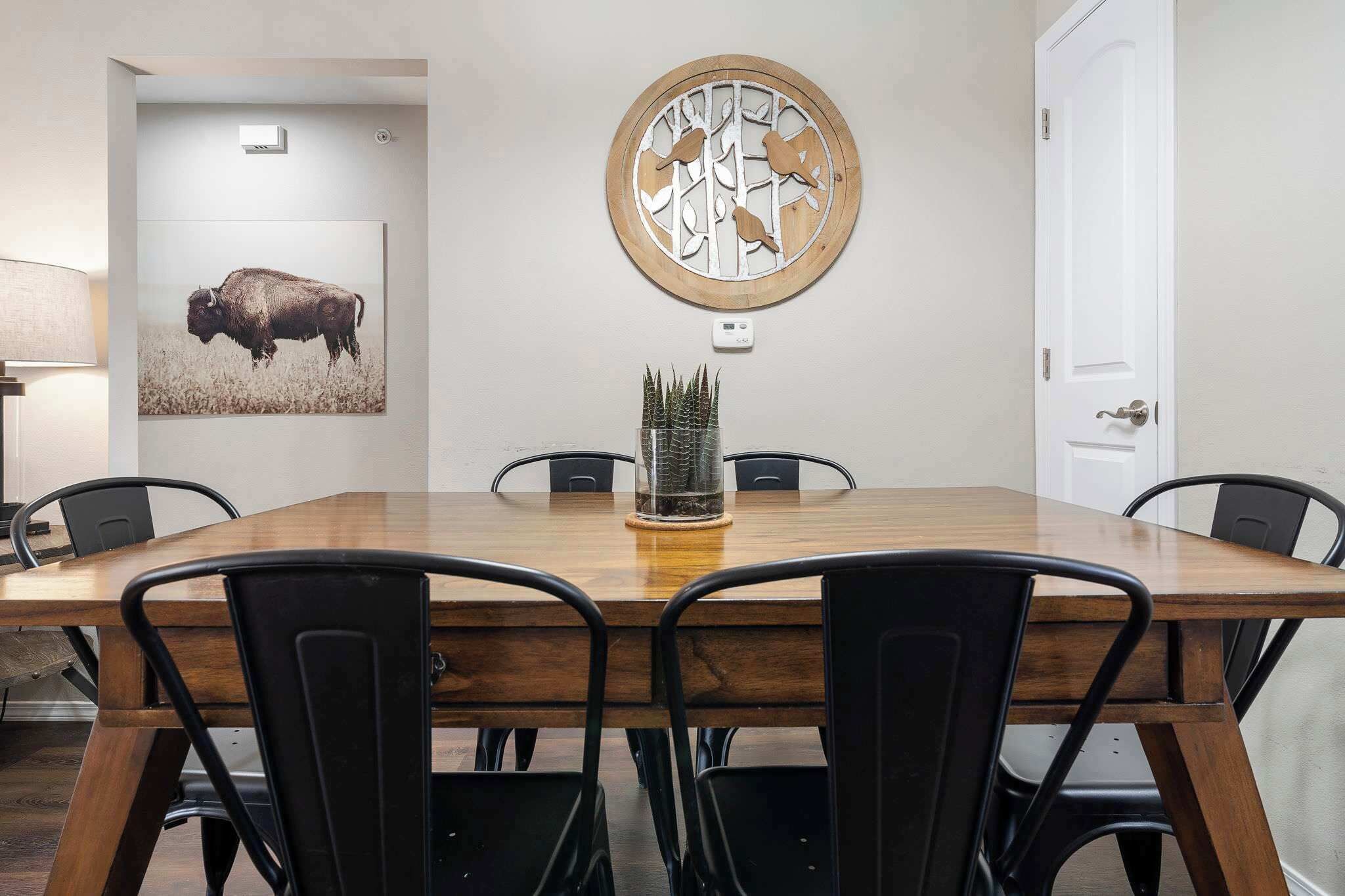 Un comedor con una mesa de madera, sillas negras, un adorno en la pared y un cuadro con la imagen de un bisonte.