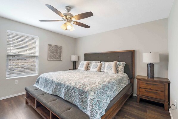 A bedroom with a bed, a bench, a ceiling fan, two bedside tables, and a window with blinds. - Urban Farmhouse - Refreshing Pool! (Branson)