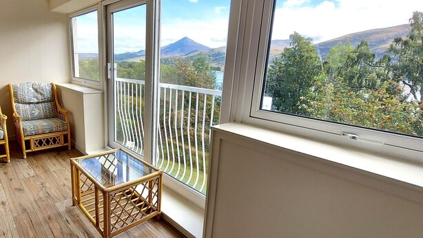 A balcony with two wicker chairs, a small table, and a view of a mountainous landscape.