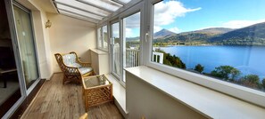 A balcony with wicker furniture, a glass table, and a view of a lake and mountains. - Otter Lodge 02 Loch Rannoch Highland Club (Pitlochry)