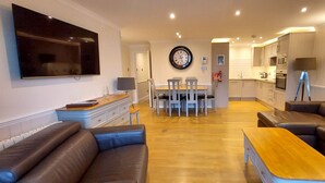 A modern living room with a large flat-screen TV, leather sofas, a wooden coffee table, and a dining area with chairs and a table. - Pine Marten Lodge 03 Loch Rannoch Highland Club (Pitlochry)