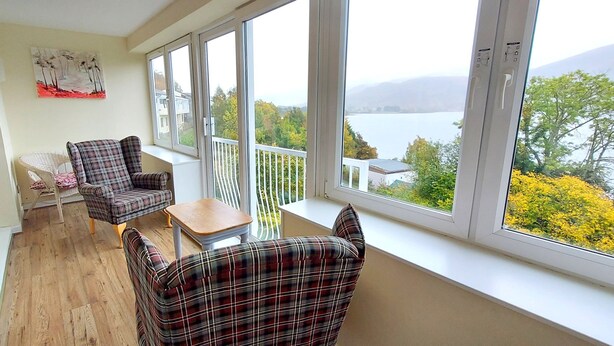 A balcony with a view of a lake and mountains, furnished with plaid chairs and a small table. - Pine Marten Lodge 03 Loch Rannoch Highland Club (Pitlochry)