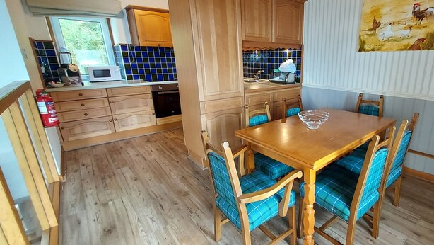 Een houten eettafel met blauw beklede stoelen in een keuken met houten kastjes en een betegelde achterwand.