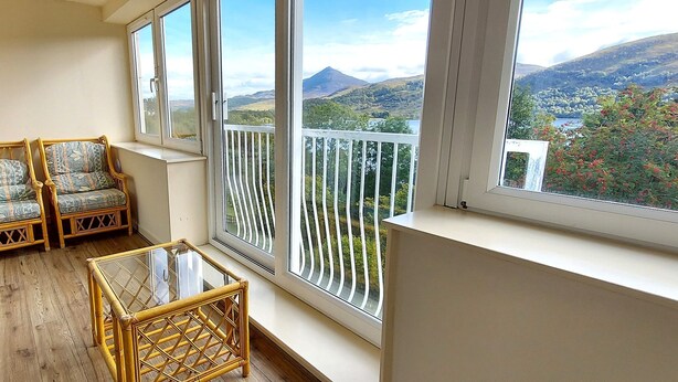 A balcony with wicker furniture, a glass table, and a view of a mountainous landscape. - Red Squirrel Lodge 05 Loch Rannoch Highland Club (Pitlochry)