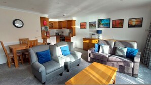 Ein Wohnzimmer mit Sofa, Sessel und Couchtisch, ein Essbereich mit Tisch und Stühlen sowie eine Küche mit Holzausbau und Geräten.
