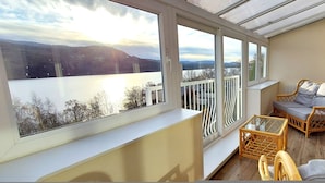 Ein Balkon mit Blick auf einen See und Berge, mit Korbmöbeln und einem Glastisch.