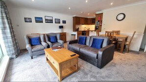 A living room with two grey sofas, a wooden coffee table, and a dining area with a wooden table and chairs. - Red Deer Lodge 05 Loch Rannoch Highland Club (Pitlochry)
