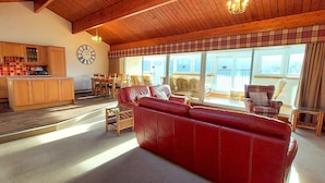 A spacious living area with a red leather sofa, wooden flooring, a kitchen with wooden cabinets, and a large window offering a view. - Highland Lodge 09 Loch Rannoch Highland Club (Pitlochry)