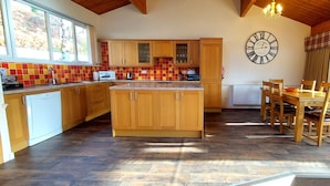 A kitchen with wooden cabinets, a tiled backsplash, a clock, and a dining area with wooden chairs and a table. - Highland Lodge 09 Loch Rannoch Highland Club (Pitlochry)