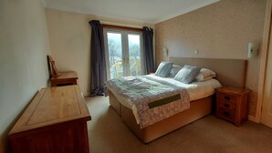 A bedroom with a bed, bedside tables, a wooden dresser, a window with curtains, and a door. - Highland Lodge 09 Loch Rannoch Highland Club (Pitlochry)