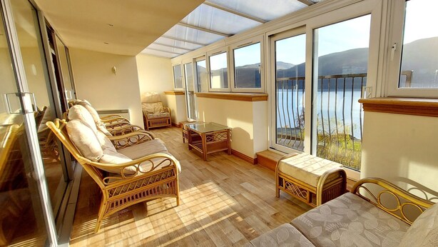 A sunlit room with wicker furniture, large windows, and a view of a lake. - Highland Lodge 09 Loch Rannoch Highland Club (Pitlochry)