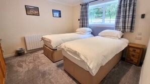A bedroom with two single beds, a window with curtains, a radiator, and a small bedside table. - Red Deer Lodge 06 Loch Rannoch Highland Club (Pitlochry)